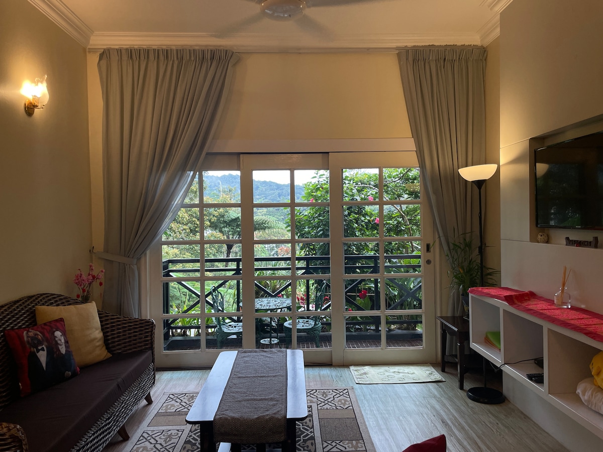 A cozy living area features stylish wicker furniture, complemented by soft throw pillows. Large glass doors provide a view of the outdoor greenery, while sheer drapes add a gentle texture. A coffee table sits in the center, with a light fixture illuminating the space.