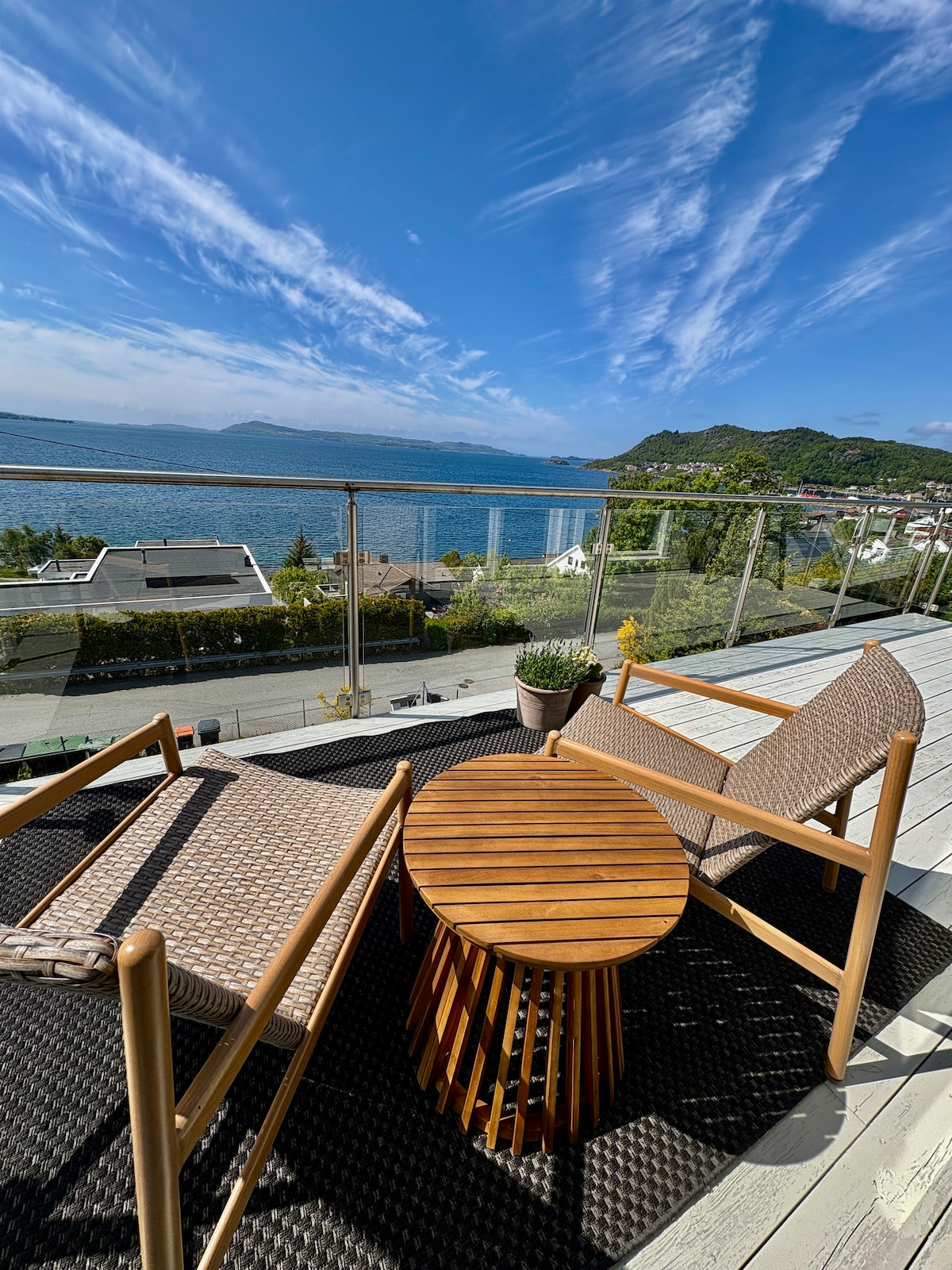 A spacious balcony is portrayed, featuring two wooden chairs and a circular wooden table. A view of the serene water and rolling hills is visible beyond the glass railing. The sky is bright with wispy clouds, enhancing the overall open feel of the space.