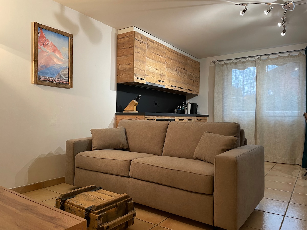 A comfortable living area features a spacious, light-brown couch, complemented by natural light filtering through sheer curtains. A wooden cabinet with a modern design is positioned above a kitchen area, and a vibrant mountain-themed artwork is displayed on the wall.