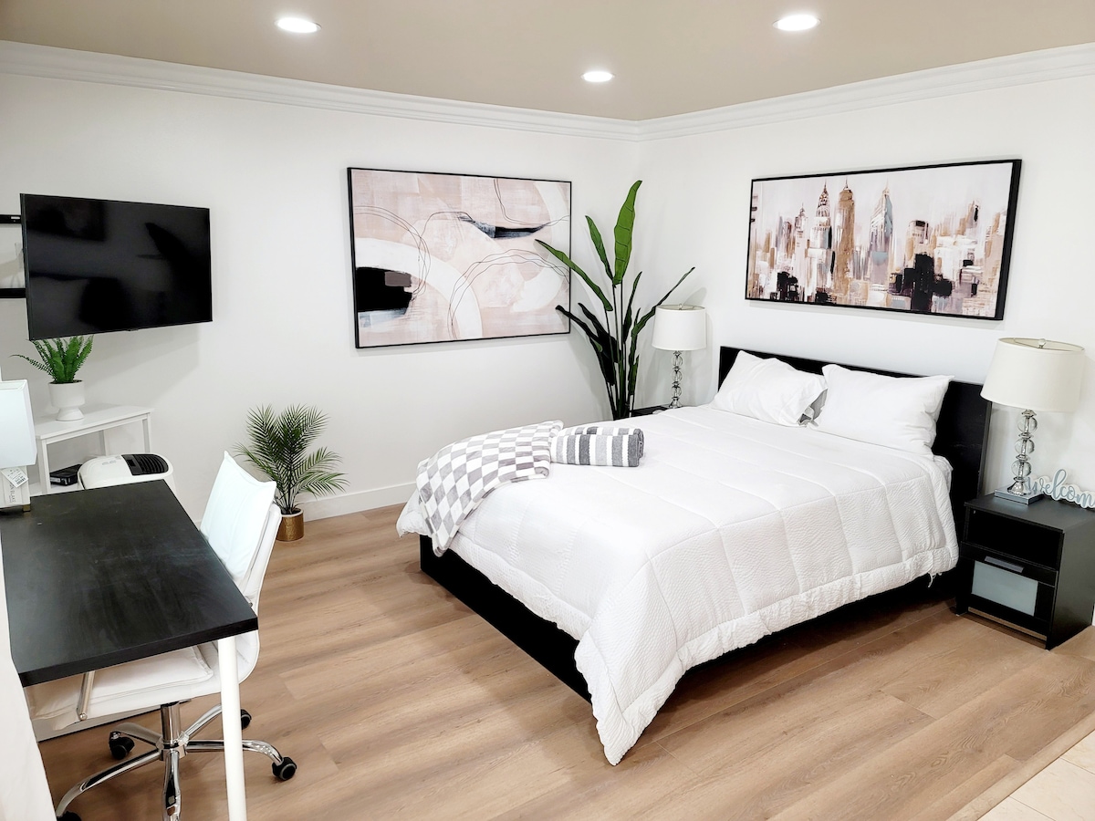 A bright studio space is featured with a spacious bed dressed in white linens, accompanied by decorative pillows. A desk and chair are positioned nearby, with a wall-mounted television above. Artwork adorns the walls, complemented by greenery from a potted plant.