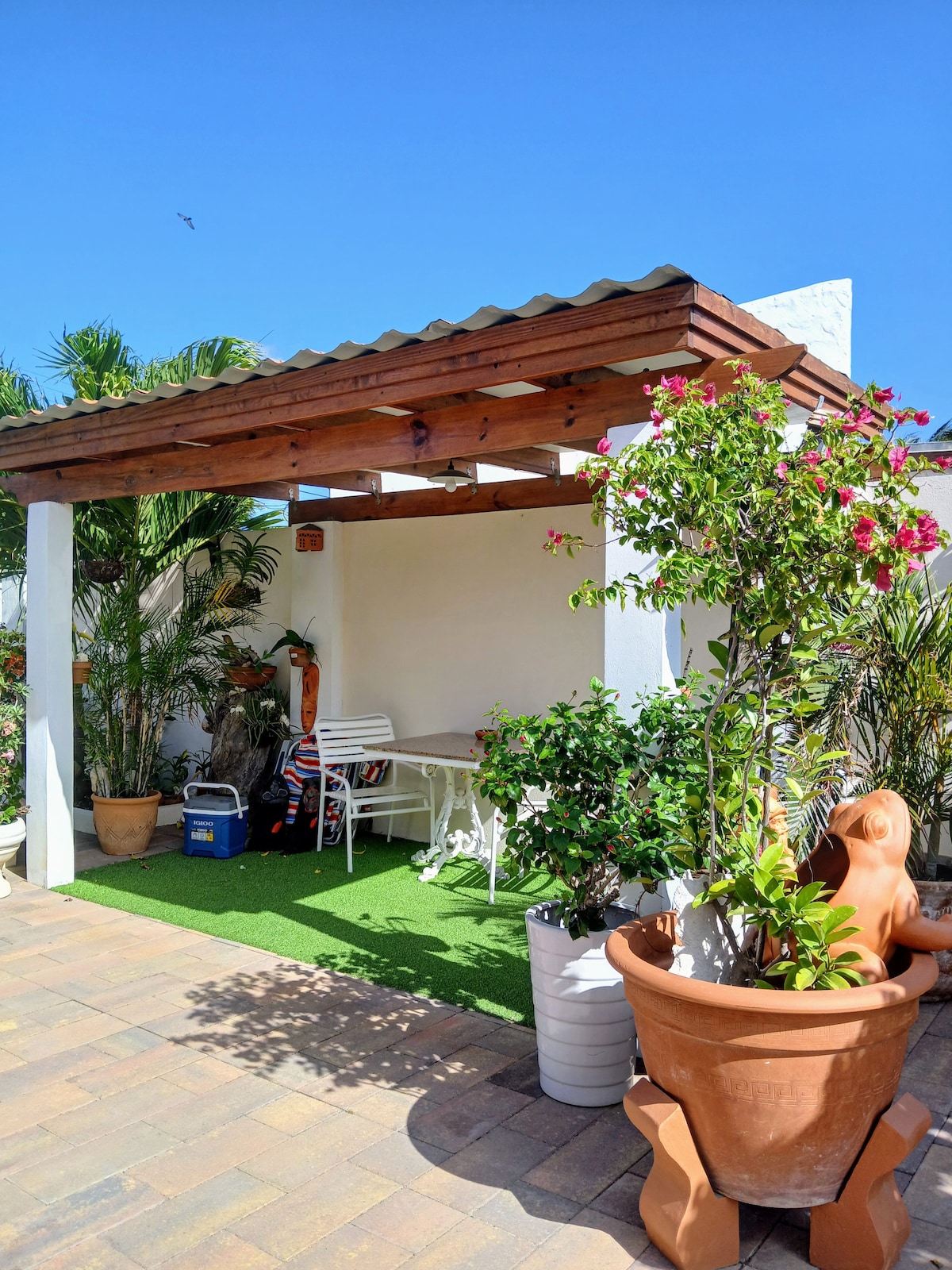 An outdoor seating area is presented under a wooden pergola, featuring a small table and chair. Lush greenery, including potted plants and colorful flowers, enhances the space. The ground is covered with green turf, and a vibrant blue sky is visible above.