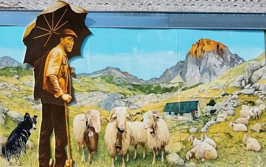 A vibrant mural depicts a figure holding an umbrella in a pastoral setting. Surrounding sheep graze on lush grass, with mountains in the background. A small cabin can be seen in the distance, enhancing the tranquil rural landscape.