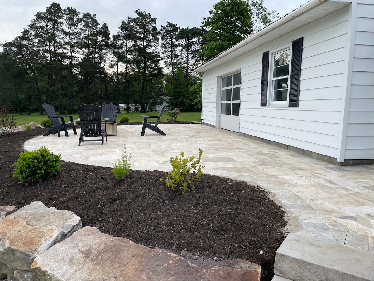 A spacious patio area features a set of black adirondack chairs surrounding a central fire pit. The patio is bordered by neatly landscaped greenery and stone edging, with a white house visible in the background. The setting offers a peaceful space for gatherings.