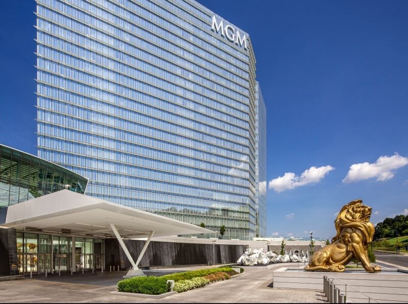 The MGM Casino building features a modern glass façade reflecting the sky. A prominent golden lion statue stands at the entrance, surrounded by landscaped greenery. The area is spacious, providing access to the entryway, creating an inviting atmosphere for visitors.