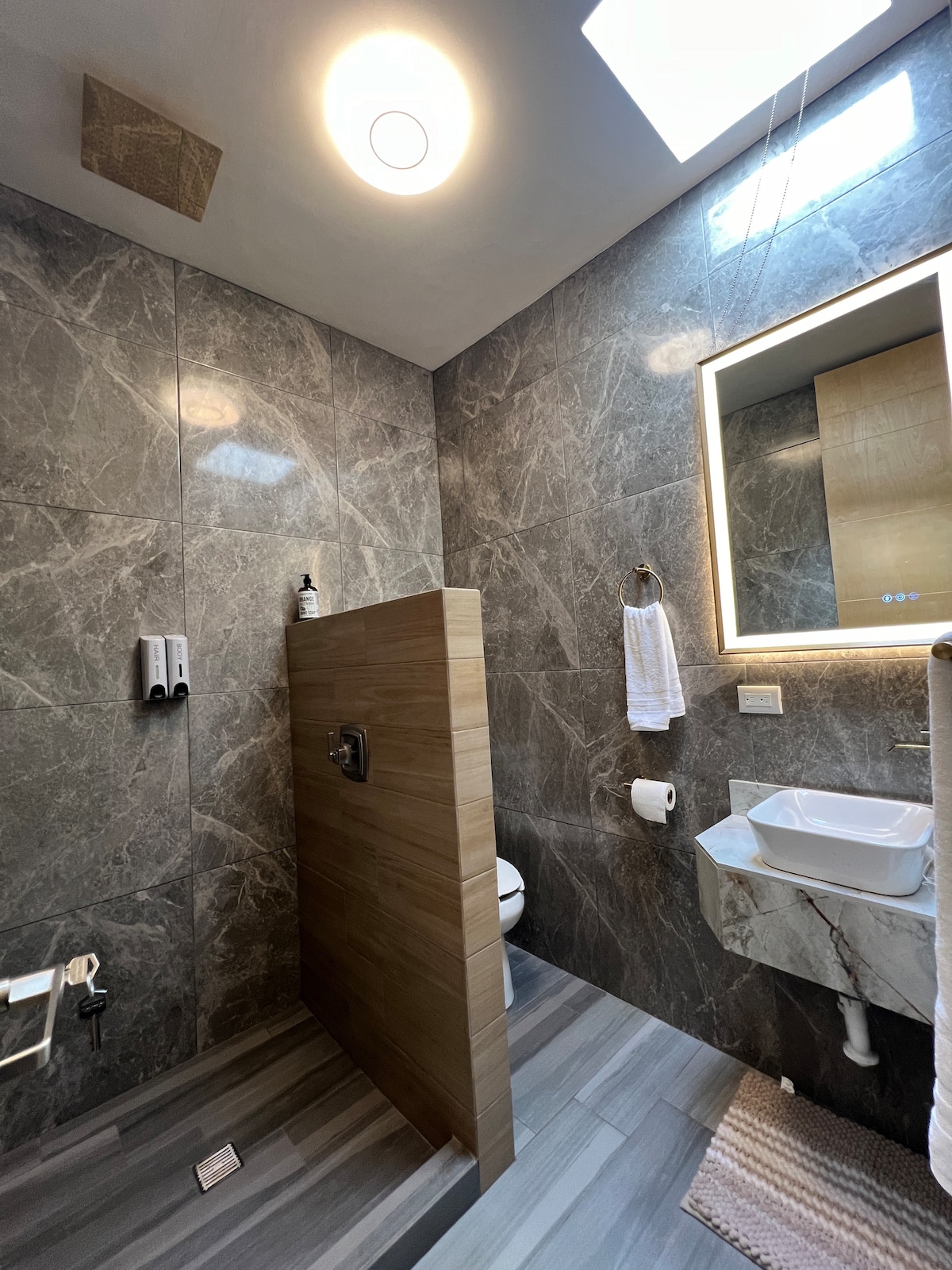 A modern bathroom features sleek marble walls and a stylish wooden partition for the shower area. A large illuminated mirror is mounted above a small sink with a marble countertop. Natural light filters in through a skylight, enhancing the overall design.