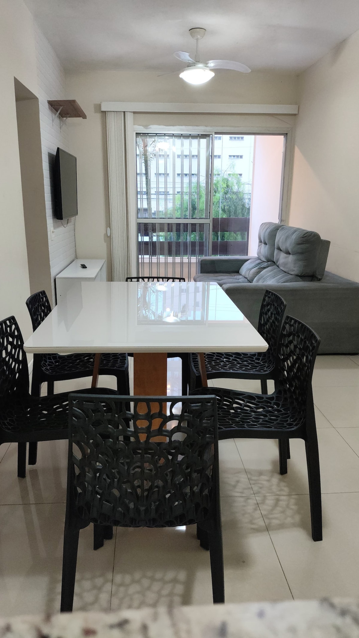 A bright and airy dining area is presented, featuring a white table surrounded by six black chairs. A cozy seating arrangement is visible in the background, with a plush gray sofa and a partially open window allowing natural light to enter.