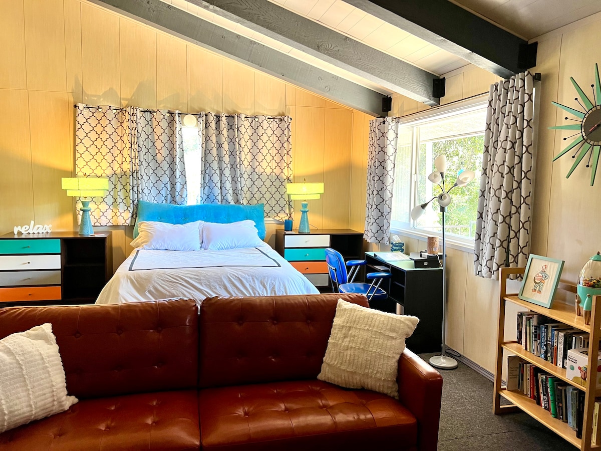 A bright room features a spacious bed with a light-colored duvet, paired with a blue accent headboard. A cozy couch occupies the foreground, while a work desk rests beside a window. Decorative lamps and art accentuate the room’s warm and inviting decor.