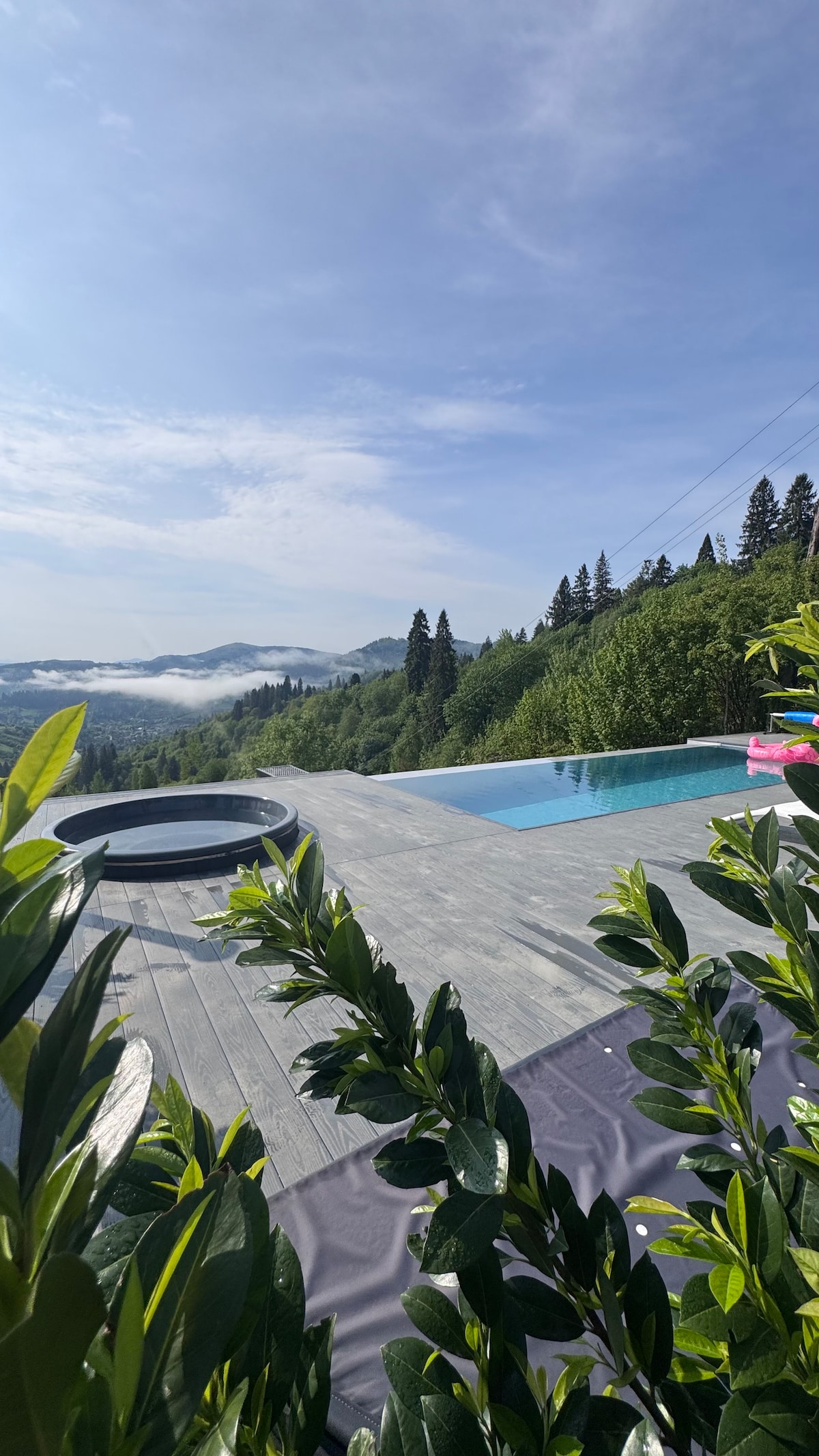 A serene outdoor area features a modern pool overlooking lush mountain landscapes. A jacuzzi is nearby on a wooden deck. Green shrubbery frames the view, adding a touch of nature to the tranquil setting. Clear skies with soft clouds enhance the picturesque scene.
