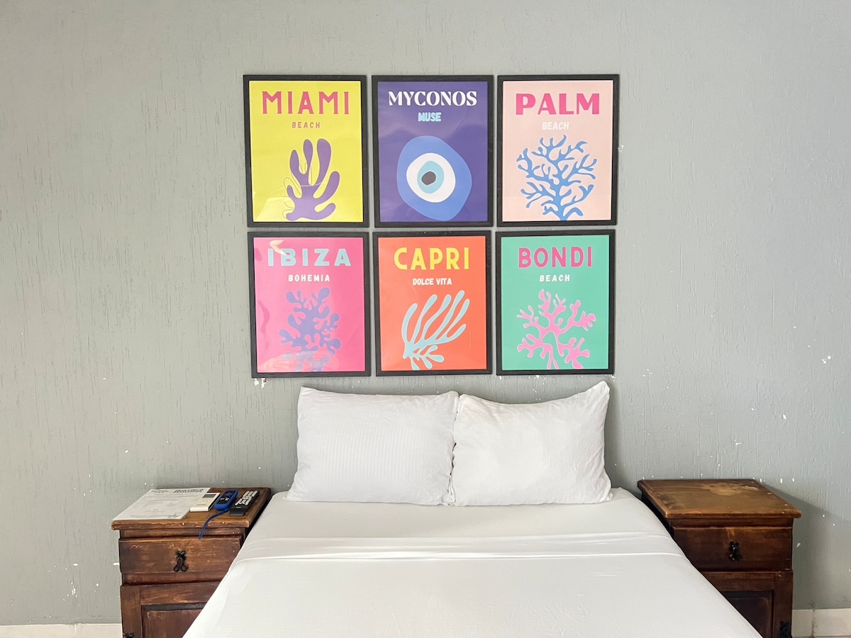 A spacious bed with white bedding is centered in front of a wall adorned with colorful framed artworks depicting various coastal destinations. The bed is flanked by wooden nightstands, adding a touch of warmth to the room's design.