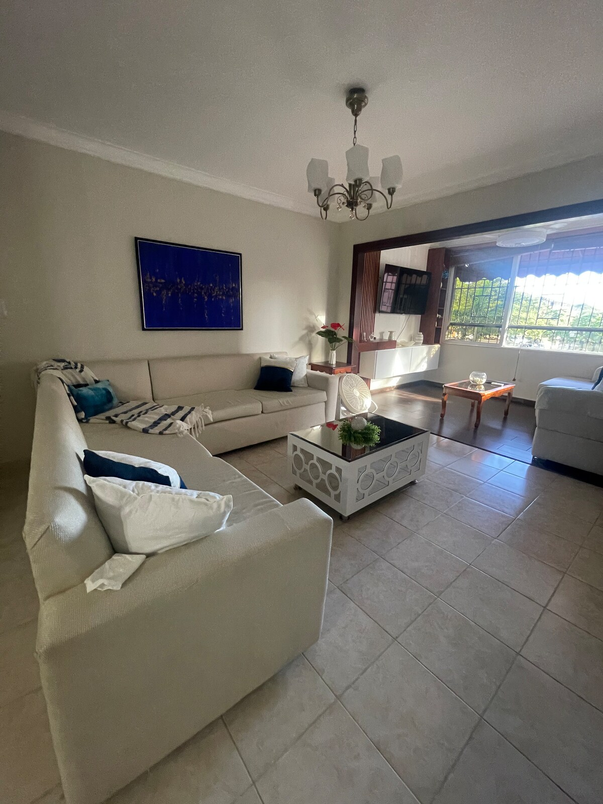 A spacious living area is arranged with light-colored sofas and a decorative coffee table centered on tiled flooring. A mural adds a touch of color to the walls. Natural light flows in from a large window, enhancing the inviting ambiance.