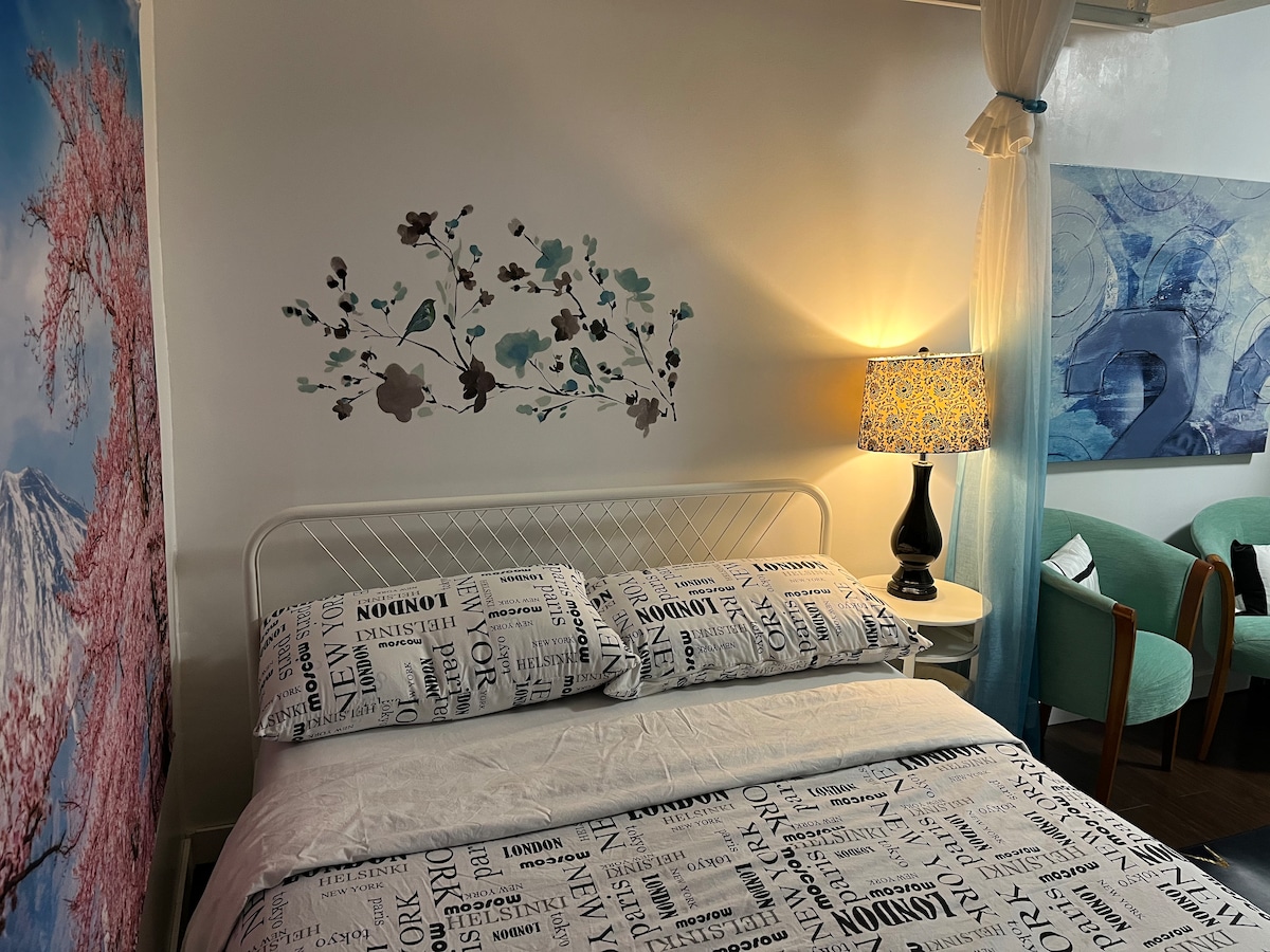A cozy sleeping area features a white metal bed with a patterned duvet cover. A bedside lamp casts a warm glow beside the bed. The wall displays floral artwork, and a small seating area is visible with light-colored armchairs. Soft drapery enhances the space.