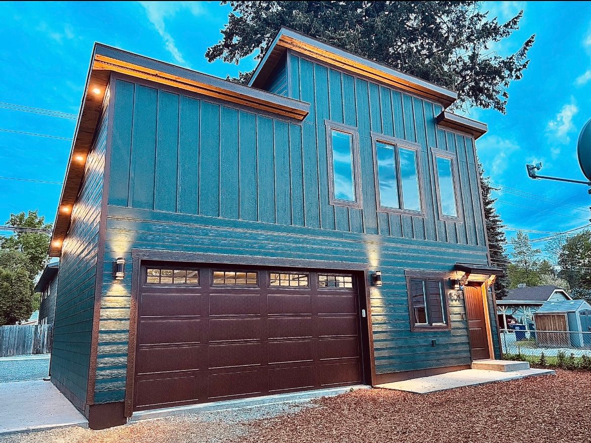 The guesthouse exterior features a modern design with a two-story structure, highlighted by dark blue wood siding and large windows. Warm outdoor lighting accentuates the architecture, while a gravel path leads to the entrance. The surrounding area includes landscaping and a clear sky.
