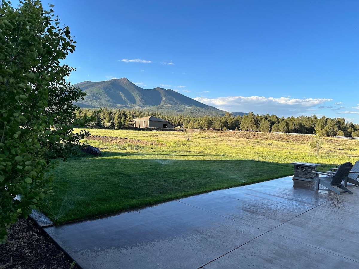 A scenic view showcases expansive green fields and distant mountains under a clear blue sky. The patio features a fire pit and outdoor seating, inviting relaxation amidst the natural landscape. Sprinklers can be seen gently watering the lawn, enhancing the tranquil outdoor setting.