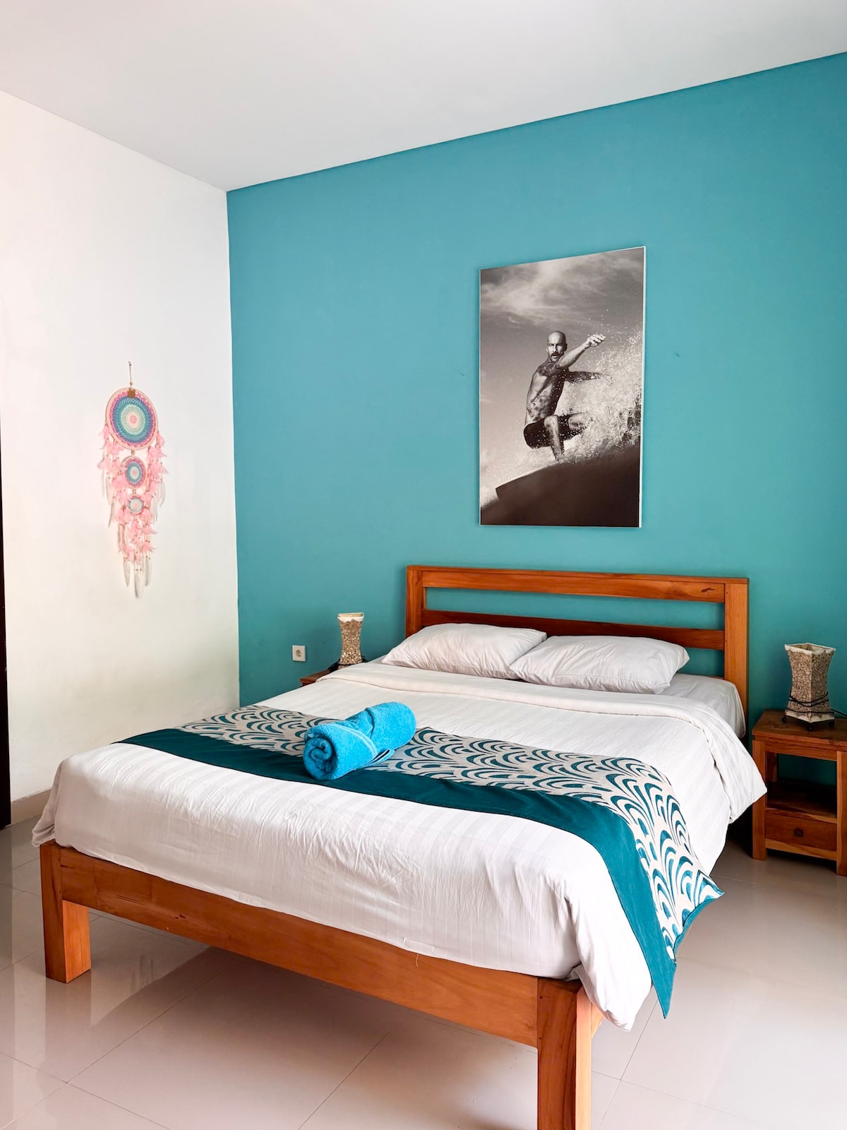 A cozy bedroom features a spacious bed adorned with white linens and a decorative throw blanket. A large photograph of a surfer is displayed on a vivid turquoise wall. Nightstands with lamps are positioned beside the bed, and a colorful wall hanging adds interest.