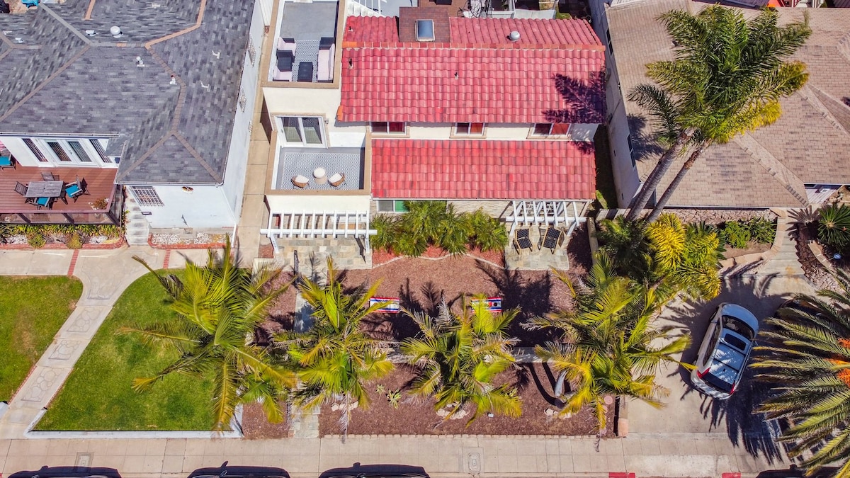 An aerial view showcases a spacious two-story home with a red-tiled roof, surrounded by well-manicured landscaping. Palm trees frame the property, while the driveway accommodates a parked car. The vibrant front yard is adorned with tropical plants, enhancing the home's coastal charm.