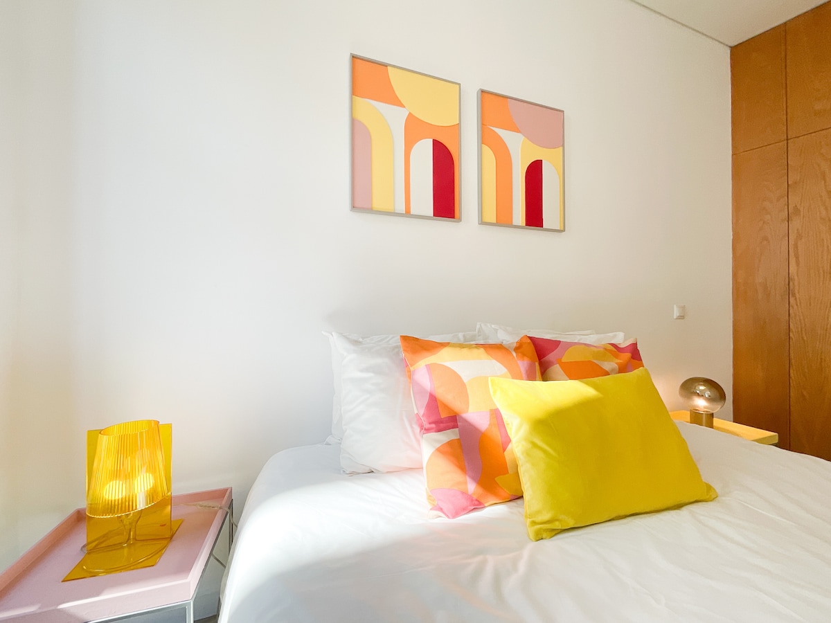 A bright bedroom features a spacious bed adorned with colorful pillows, complemented by a vibrant yellow cushion. Two abstract art pieces are displayed above the bed, while a bedside table holds a decorative lamp. Natural light illuminates the room, enhancing its welcoming ambiance.