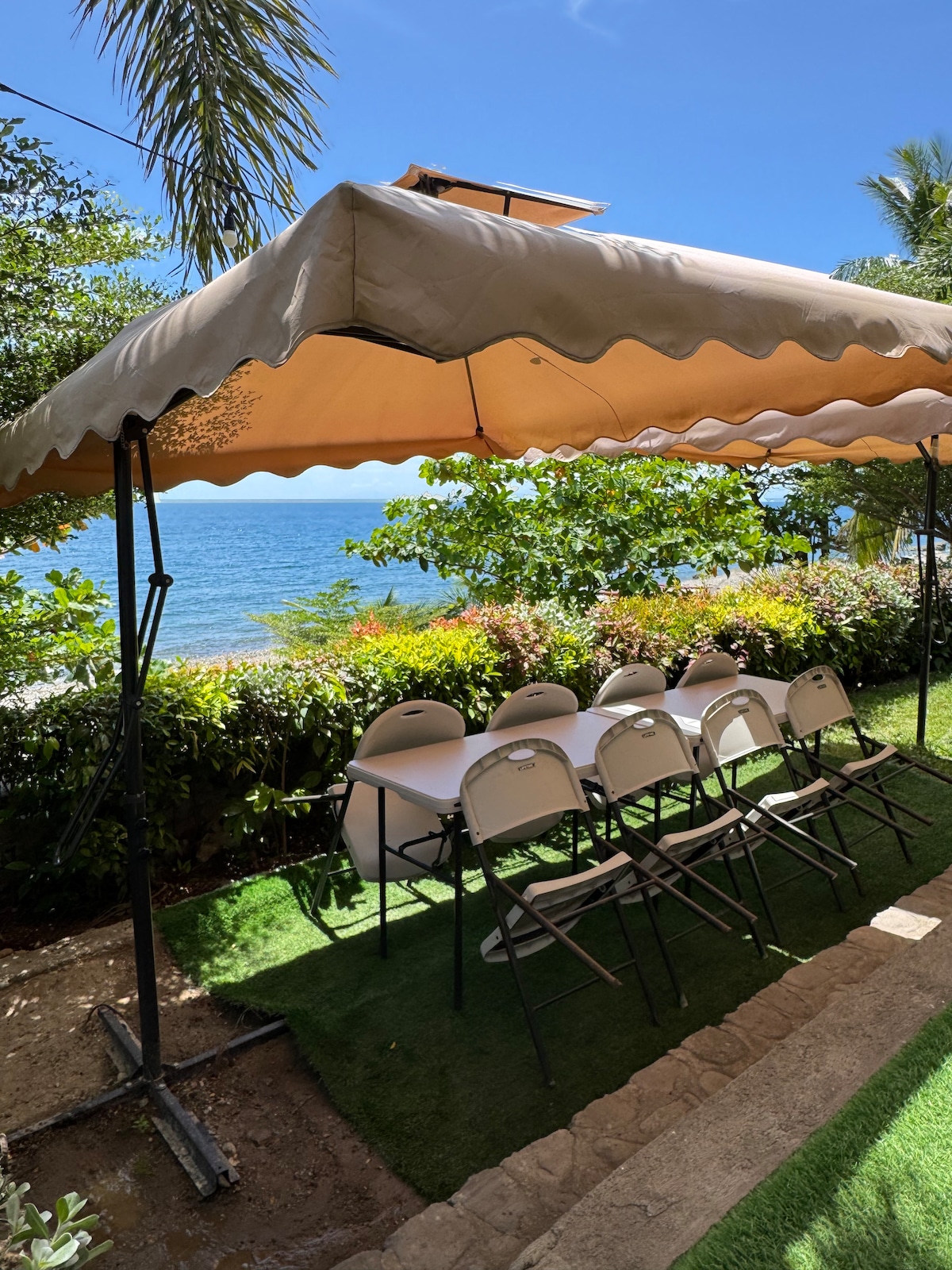 A shaded outdoor seating area is presented with a large canopy, overlooking the ocean. Several light-colored folding chairs surround a rectangular table, with greenery framing the space. Sunlight filters through the leaves, creating a serene atmosphere for gatherings.