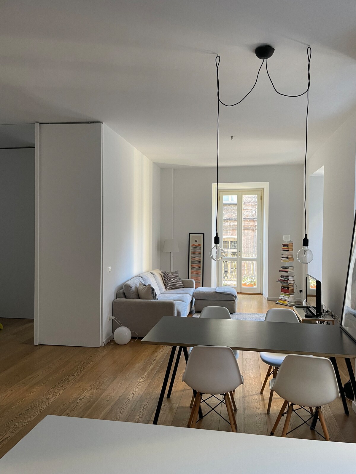 An open space design features a light-filled living area with a comfortable sofa and a dining table surrounded by several white chairs. Large windows provide natural light, and minimalist decor emphasizes the clean lines and modern aesthetic of the apartment.