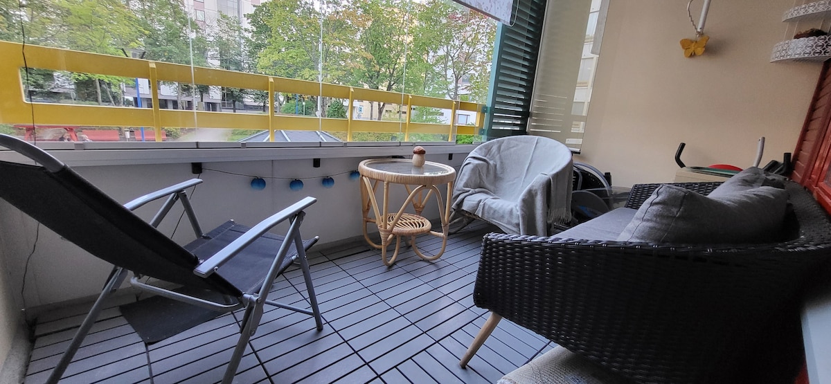 A spacious glass-enclosed balcony features comfortable seating consisting of a wicker couch and a lounge chair. A round table is positioned at the center, surrounded by green foliage visible through the glass. The floor is made of wooden decking, providing a warm aesthetic.