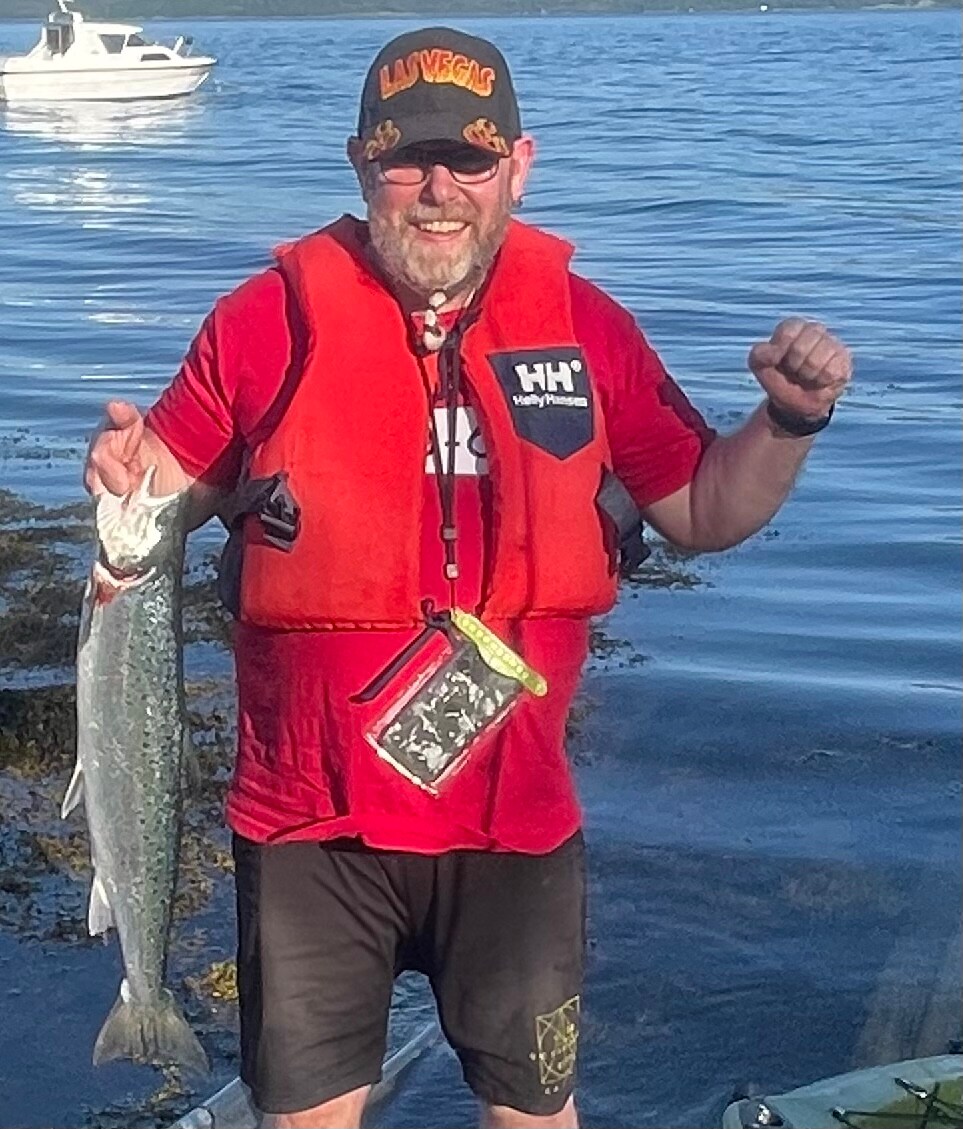 A person wearing a bright orange life vest holds a fresh fish in front of them. The background features calm water with a boat visible in the distance. The scene is set in a natural environment, emphasizing the outdoor fishing experience.