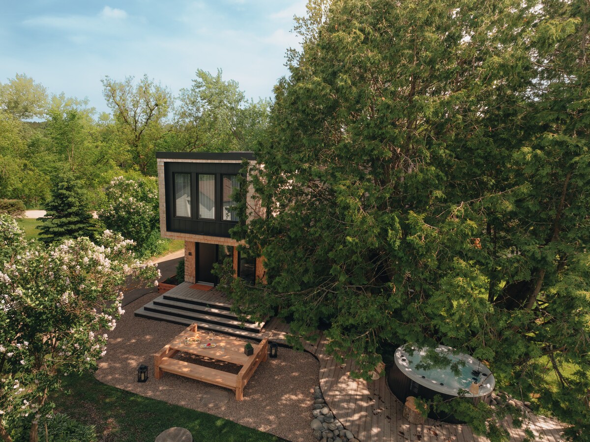 The view captures a modern chalet surrounded by greenery, featuring a spacious entrance with stone steps leading to large windows. A jacuzzi is visible to the right, nestled under a large tree, with a wooden deck and outdoor seating area in the foreground.