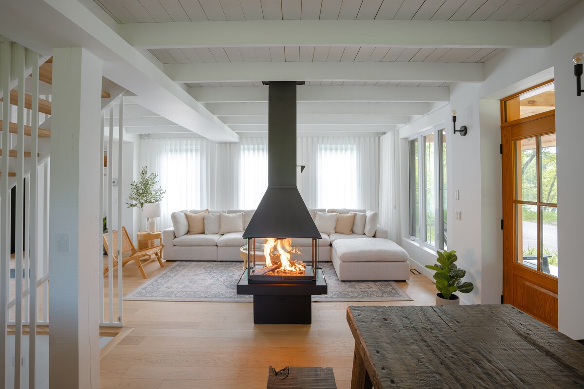 A spacious living area is highlighted by a central four-sided gas fireplace, surrounded by a comfortable seating arrangement. Large windows are adorned with light sheer curtains, allowing natural light to fill the room. A wooden table and greenery add to the inviting atmosphere.