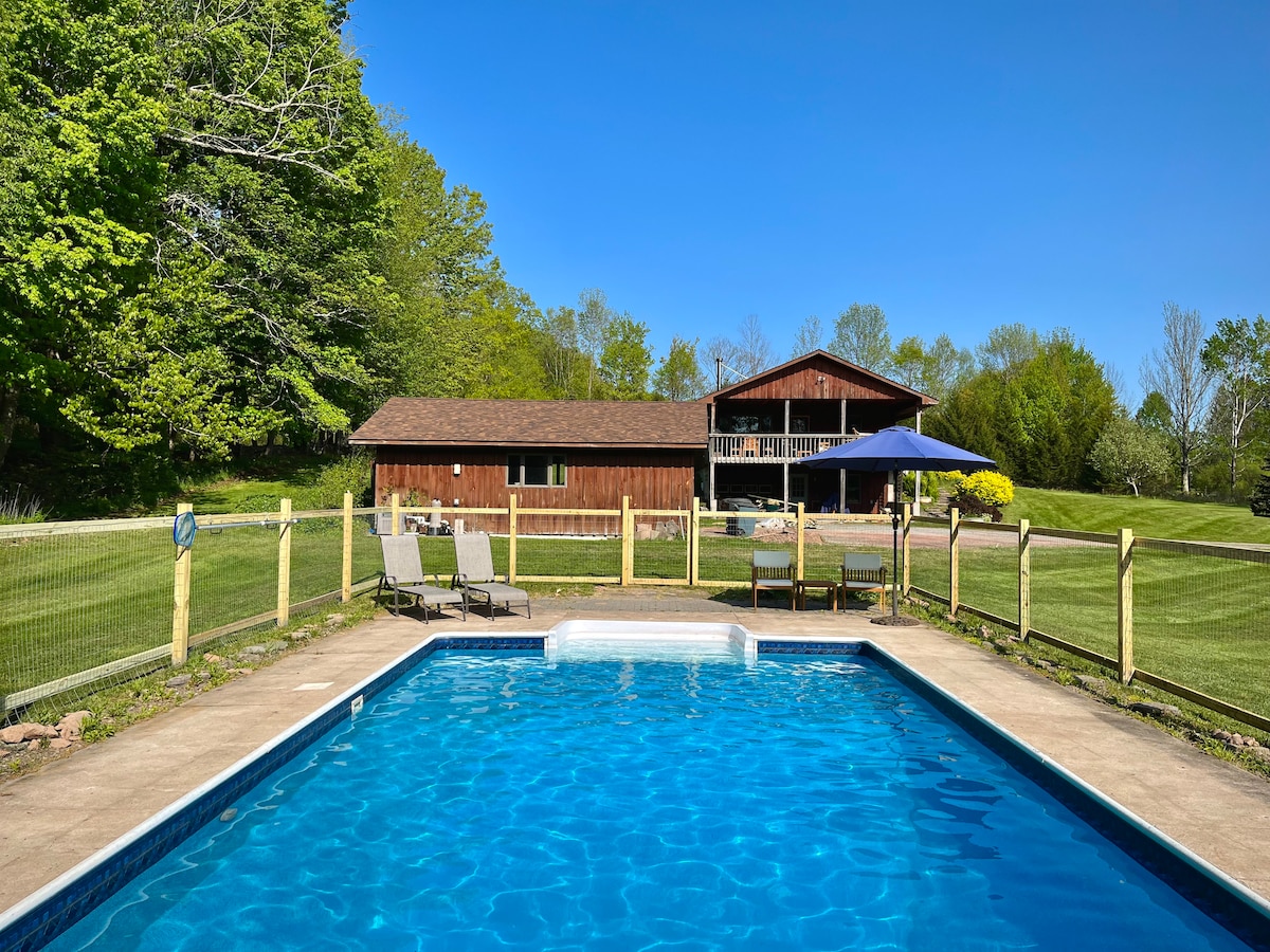 A clear blue pool is framed by a spacious lawn, with comfortable chaise lounges positioned nearby under an umbrella. In the background, a rustic cabin is set against lush greenery and a bright blue sky, creating a serene outdoor environment.