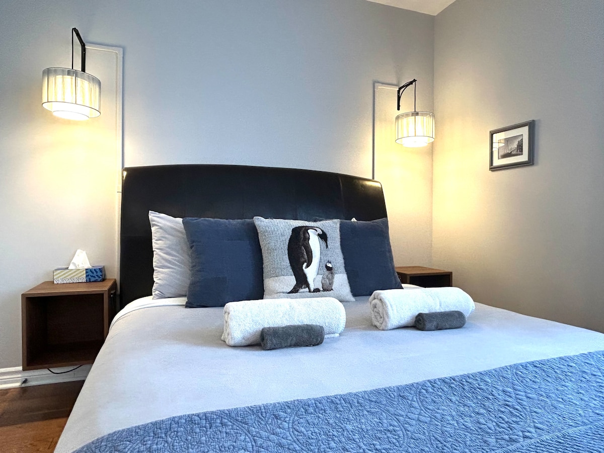 A spacious bedroom is highlighted by a large bed with decorative pillows and neatly arranged towels. Two bedside lamps provide soft lighting, while a piece of wall art adds subtle detail. Dark wooden nightstands are positioned on either side of the bed.