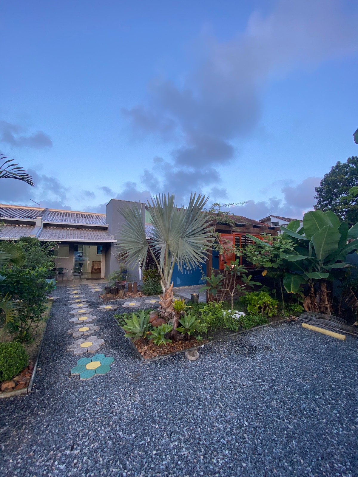 A garden area is showcased, featuring a variety of plants, including palm trees and lush greenery. A pathway made of colorful stones winds through the garden, leading to the entrance of the flat, with the evening sky visible above.