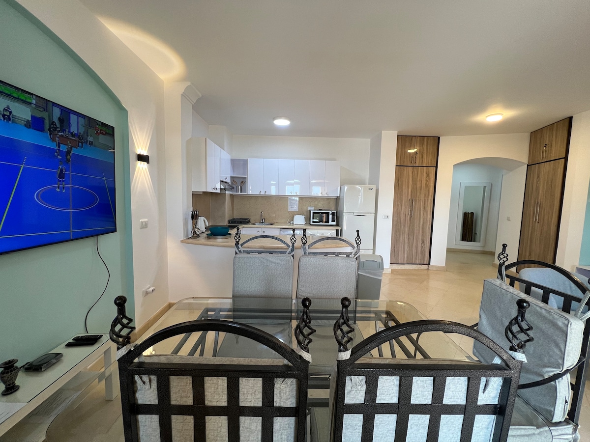 A modern open-plan kitchen and dining area is featured, with a large dining table surrounded by stylish black chairs. A wall-mounted TV displays a sports broadcast, while kitchen appliances are visible, enhancing functionality in the bright space with marble flooring.