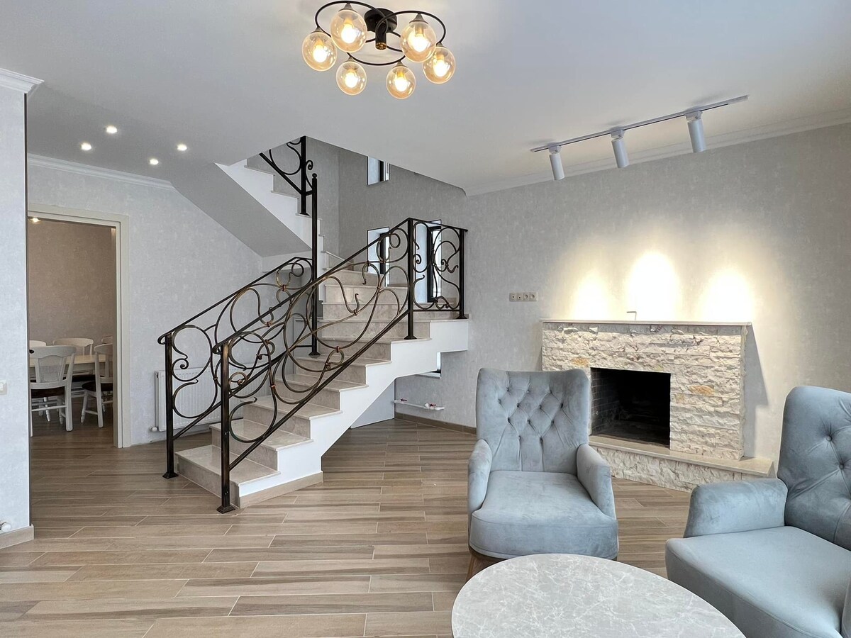 An inviting living space features a staircase with elegant railings and a modern chandelier overhead. A stone fireplace serves as a central focal point, while two plush armchairs are arranged around a light-colored coffee table on a warm wooden floor.