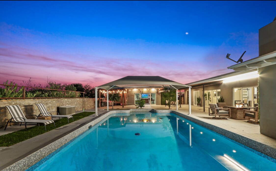 A serene evening setting is displayed, featuring a clear pool reflecting the vibrant colors of the twilight sky. A shaded seating area is visible beside the pool, framed by lush greenery and cozy lighting that enhances the outdoor space.