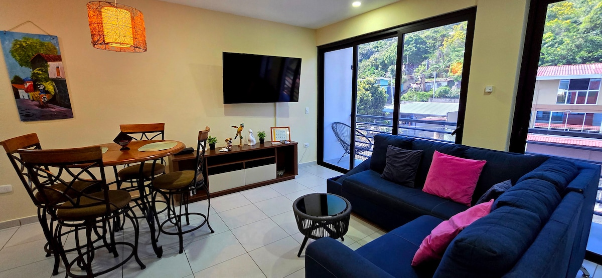 A modern living area is presented with a dark blue sofa accented by colorful cushions. A wooden dining table with four chairs is visible, alongside a wall-mounted flat-screen TV. Large glass doors frame the view, allowing natural light to fill the space.