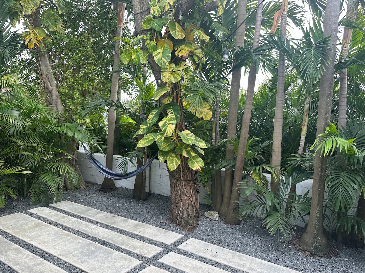 A tropical garden is visible, featuring a variety of lush green plants and palm trees. A hammock is hung between trees, offering a relaxing spot. Stone pathways lead through the garden, bordered by gravel for easy navigation.