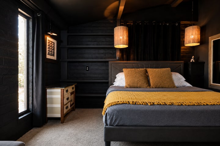 Cozy king bedroom with dramatic charcoal walls, warm pendant lighting and layered neutral bedding. Tan pillows and a mustard-yellow throw add warmth to the room’s distinctive desert-modern style.
