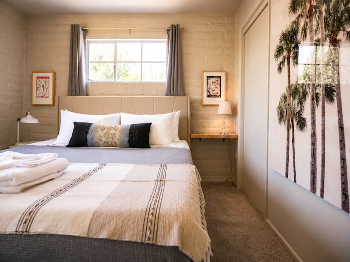 Wake up to Tucson sunshine in this cozy, art-filled bedroom. Soft neutral bedding, warm bedside lighting, and oversized palm tree art create a relaxed desert retreat with plenty of personality.