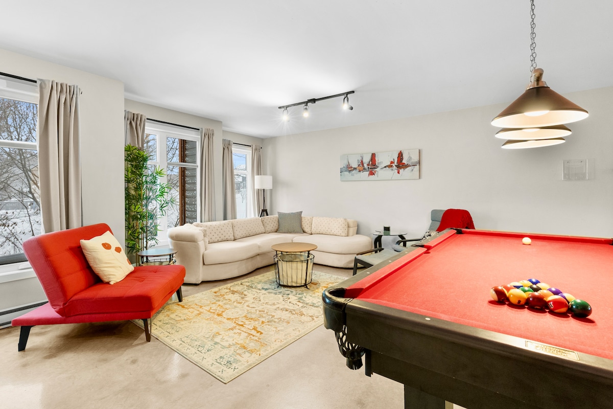 The living area features a pool table in the foreground, with a red felt surface and colorful balls arranged. A light-colored couch and an accent chair in bold red provide comfortable seating, while art hangs above a casual coffee table. Large windows allow natural light to brighten the space.