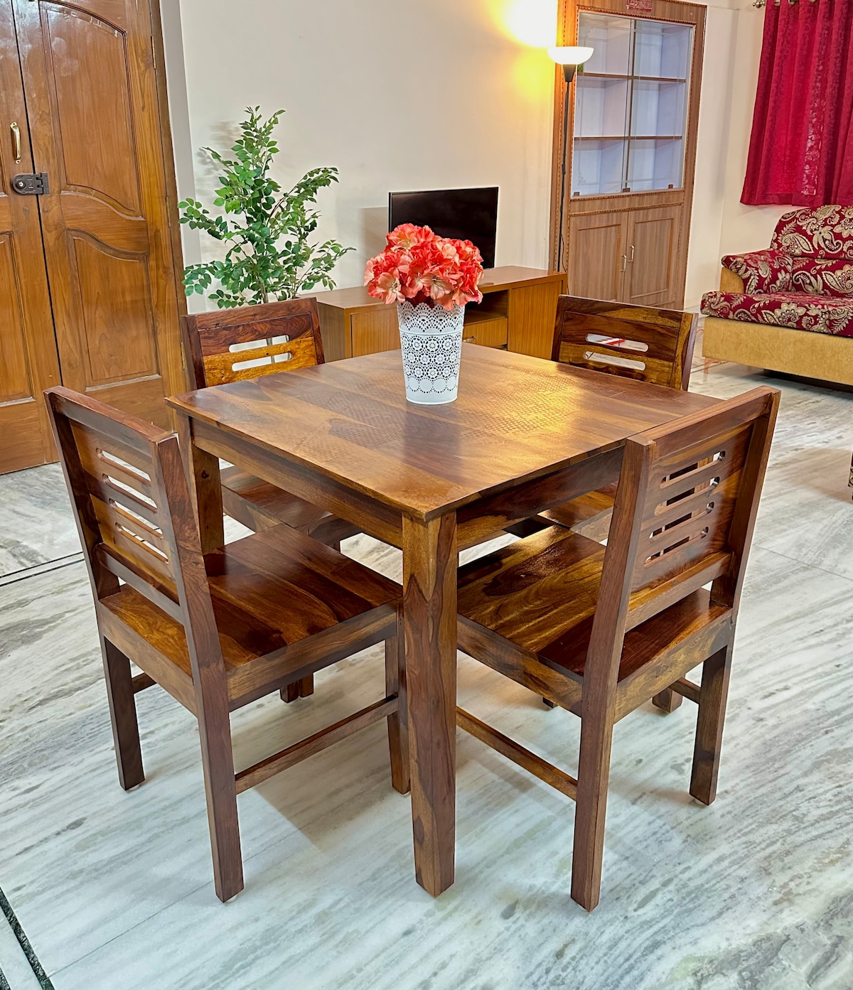 A wooden dining table is surrounded by four matching chairs, creating a cozy dining area. A vase adorned with flowers sits center stage on the table. Warm lighting enhances the inviting feel of the space, with a subtle backdrop of decorative curtains and furniture.