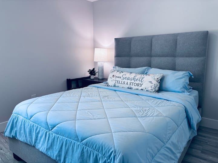 Dhomë gjumi komode me një krevat dopio "queen" me kokë krevati të veshur me susta moderne, shtroja të buta blu dhe detaje bregdetare. Një abazhur pranë krevatit dhe një komodinë shtojnë ngrohtësi, duke krijuar një ambient relaksues për net të qeta pranë plazhit.