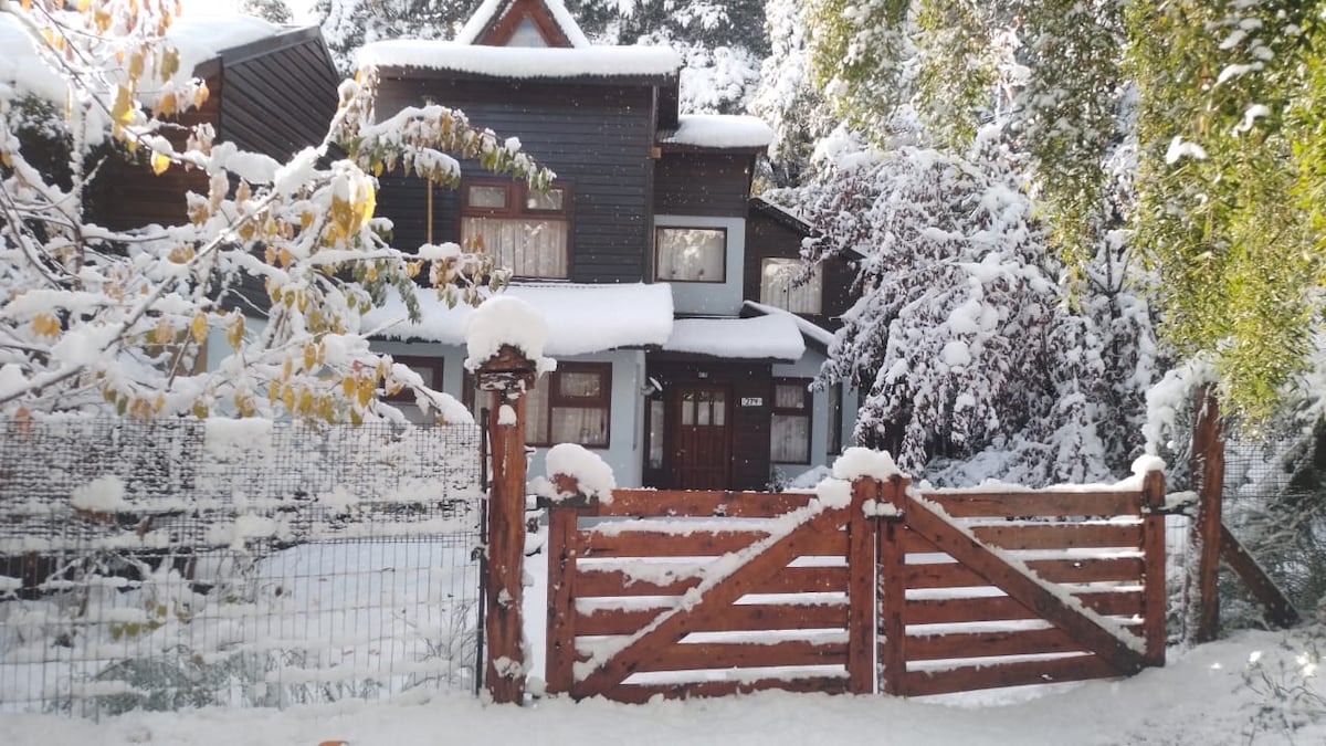 A two-story house is surrounded by a blanket of fresh snow, creating a serene winter landscape. The wooden gate leads to a warm entrance, while trees and shrubs are covered in snow, enhancing the calm atmosphere of the setting.