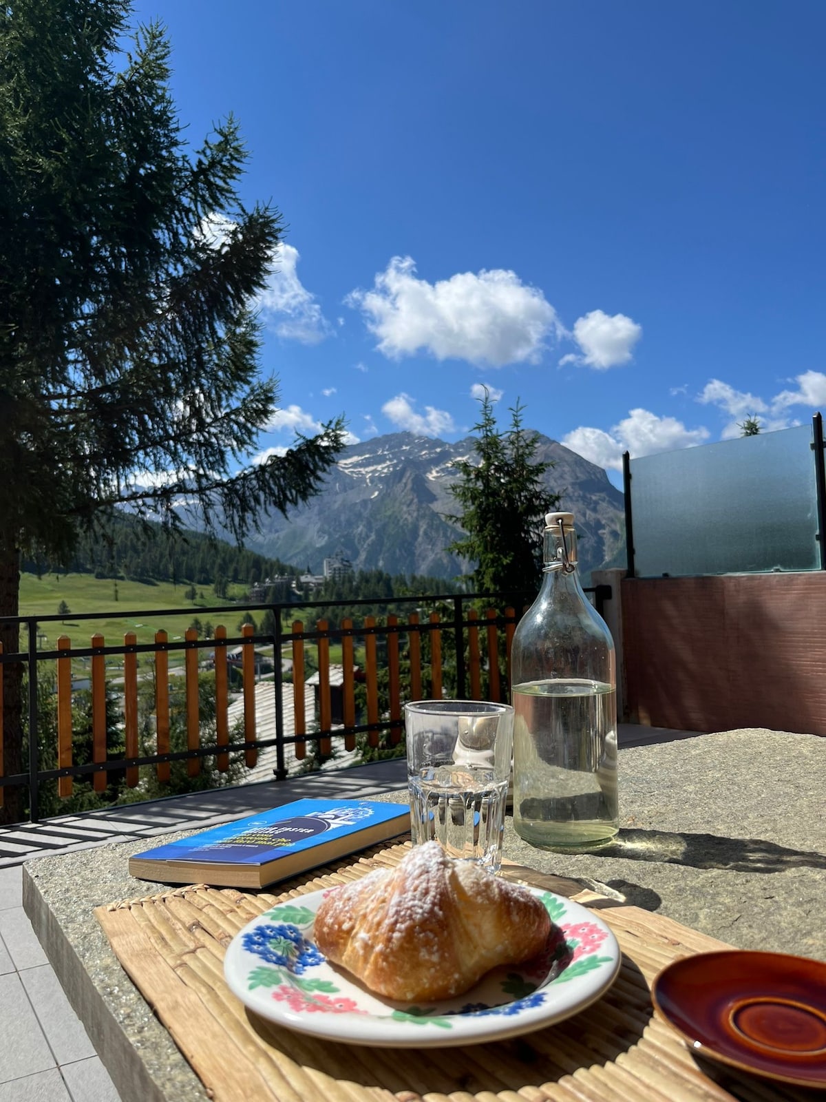 A terrace table is set with a glass of water and a croissant on a floral plate, surrounded by greenery and mountains in the background. A book rests on the table, and the sky is bright with scattered clouds.