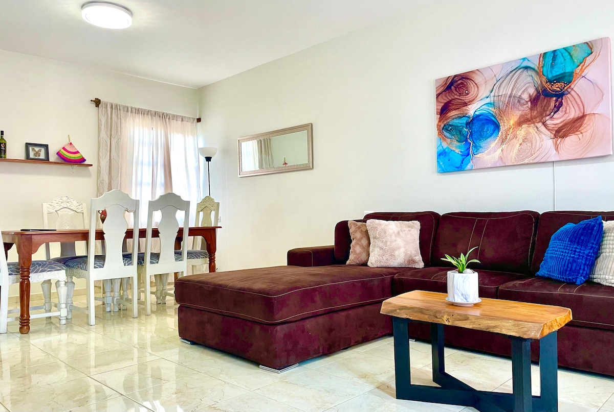 A spacious living area showcases a large sectional sofa in a rich burgundy hue, complemented by decorative pillows. A wooden coffee table sits in front, while a dining area with white chairs and a table is visible in the background. Natural light filters through a nearby window.