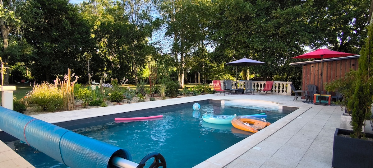 A swimming pool is surrounded by a landscaped garden with lush greenery. Inflatable toys float in the water. Loungers and umbrellas provide shaded seating areas, and a patio with outdoor furniture is visible under a canopy of trees.