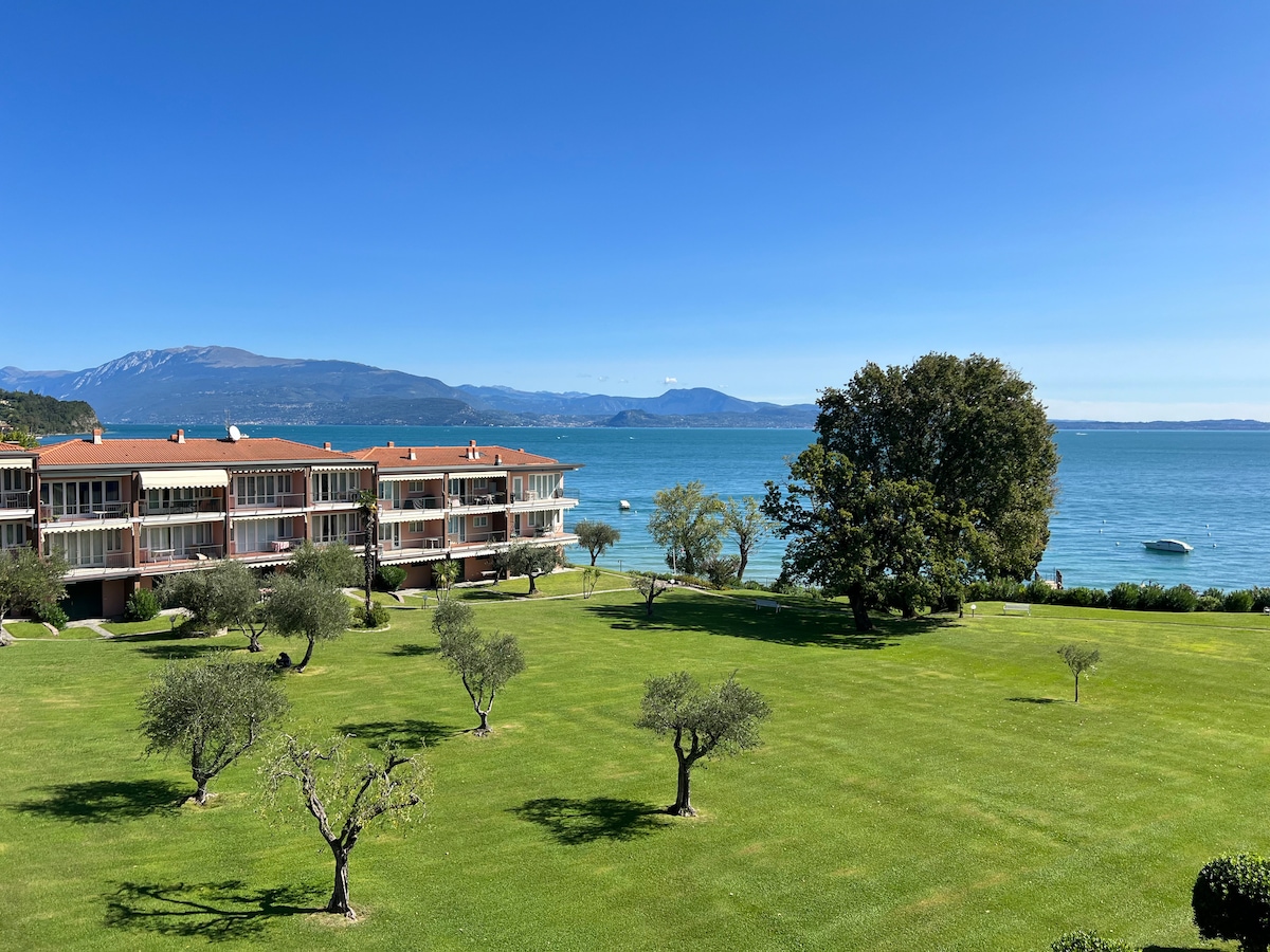 The image captures a serene view of a manicured garden featuring olive trees and a well-maintained lawn. The apartment complex is visible in the background, with a clear view of the lake and distant mountains under a blue sky.