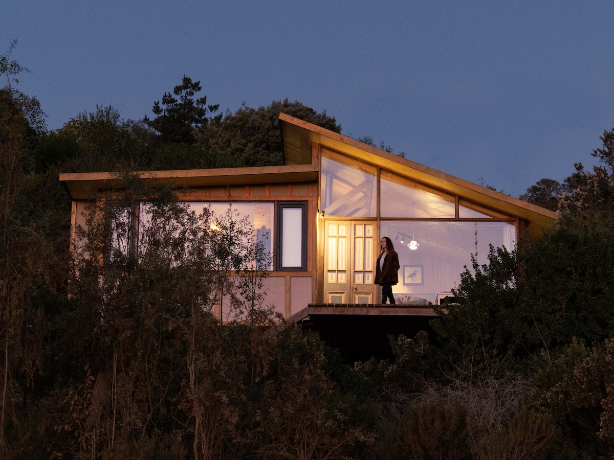 A wooden house with a modern design is shown at dusk, featuring large windows that illuminate the interior warmly. Surrounded by greenery, the structure blends with the landscape, highlighting the spacious entryway and inviting ambiance.