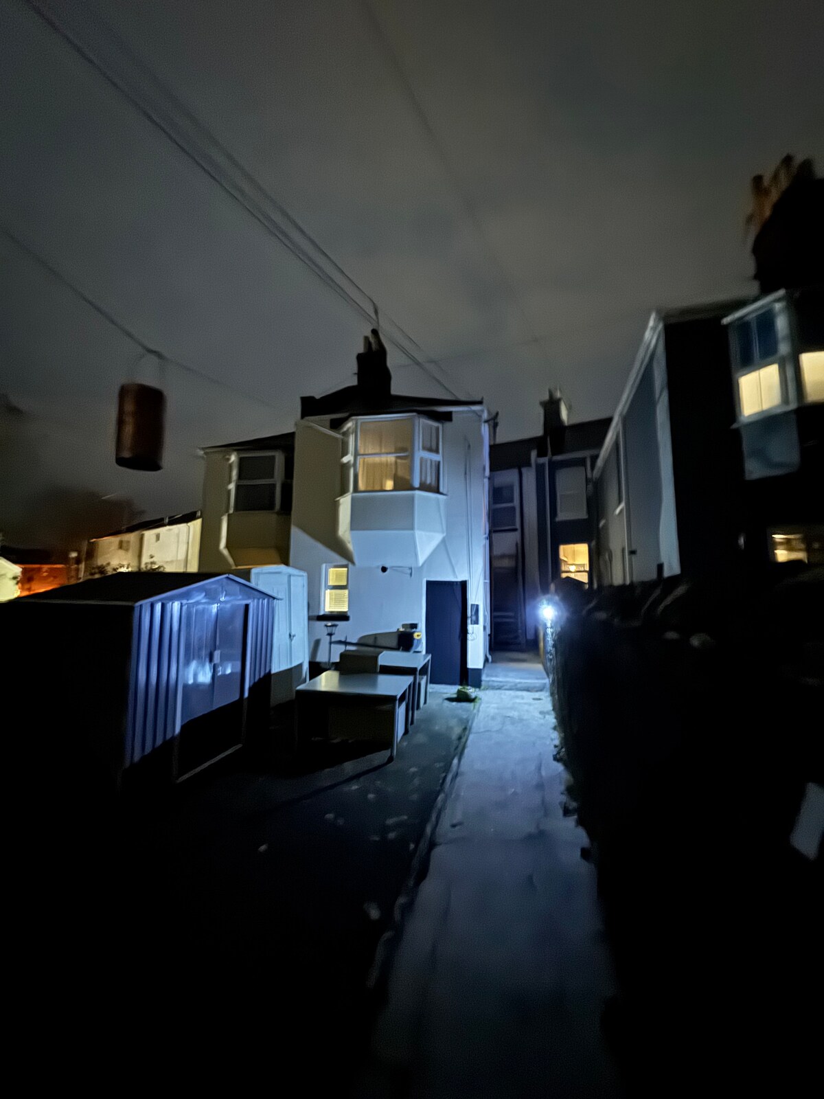 A view of the rear of the property is presented during the night. The structure features a two-story design with illuminated windows. A pathway runs alongside the building, leading to a small outdoor seating area surrounded by a fence. Nearby structures are visible in the dim light.