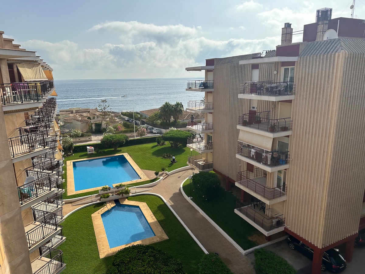 A view of the residential area showcases two swimming pools surrounded by landscaped gardens. Apartments with balconies overlook the pools and the sea beyond, while paths connect the spaces. The sky is partly cloudy, suggesting a pleasant atmosphere.