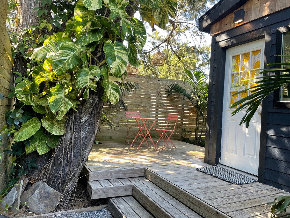 A private deck space features two pink chairs and a small table, nestled among lush greenery. The entrance to the studio is visible, framed by tropical plants and wooden slats, which enhance the outdoor tranquility.
