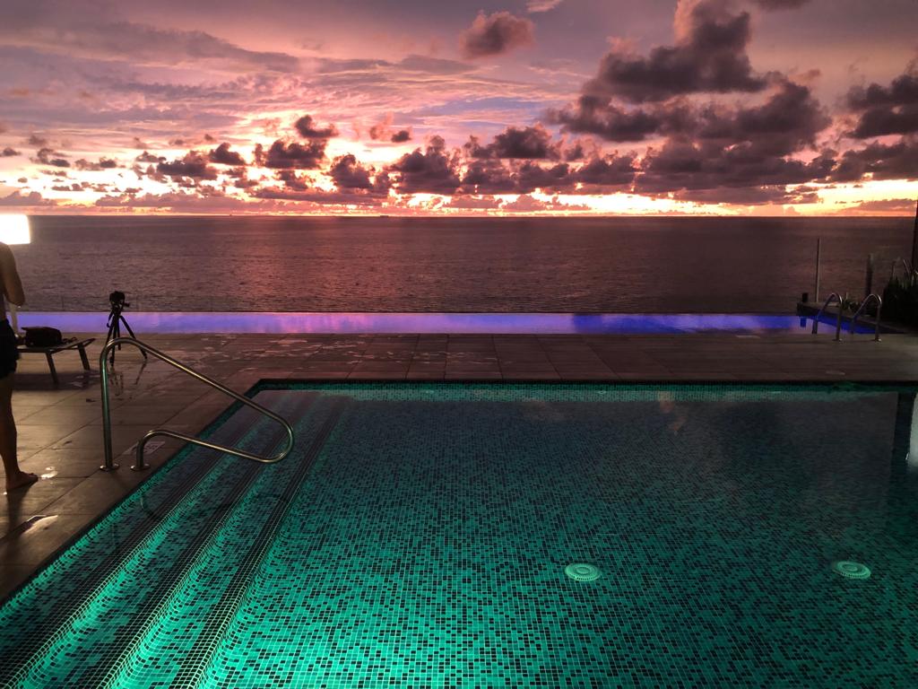 A serene infinity pool reflects the vibrant colors of a sunset, with hues of pink, orange, and purple in the sky. The pool's edge blends seamlessly into the ocean. Surrounding the pool, tiles are arranged in a mosaic pattern, enhancing the tranquil ambiance.