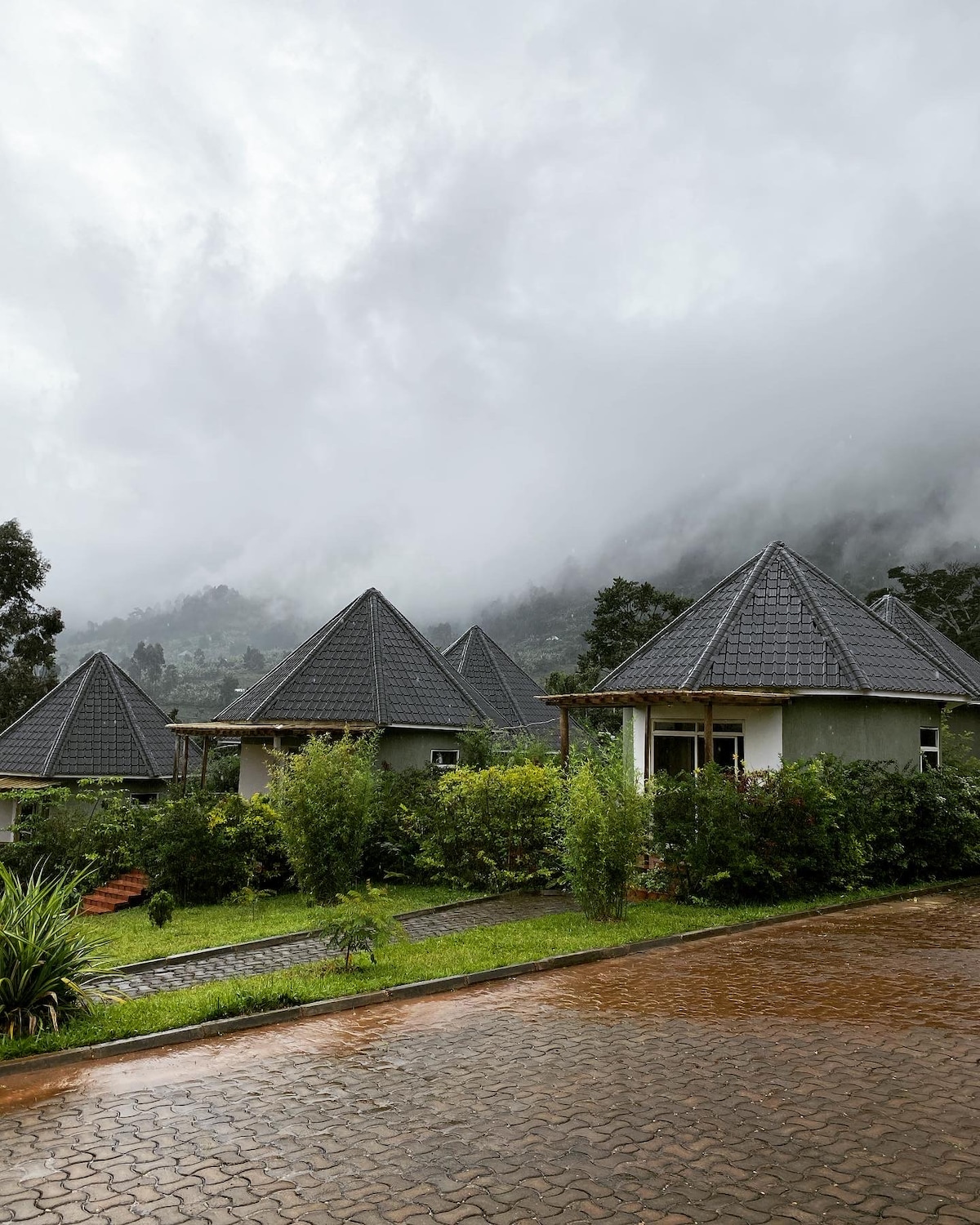 Several independent cottages are nestled among lush greenery under a cloudy sky. The cottages feature distinctive triangular roofs and are spaced for privacy. A neat pathway leads up to a cottage entrance, with a backdrop of misty mountains partially obscured by clouds.
